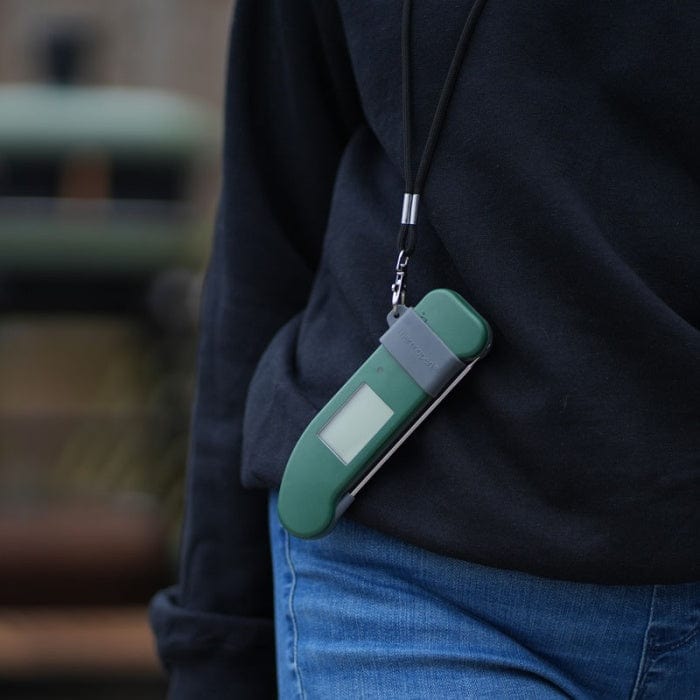 Une personne portant un jean et un sweat-shirt foncé porte un appareil numérique vert autour du cou sur une sangle, un peu comme la commodité de la Bande magnétique Thermapen avec cordon de Thermometre.fr pour un accès rapide.