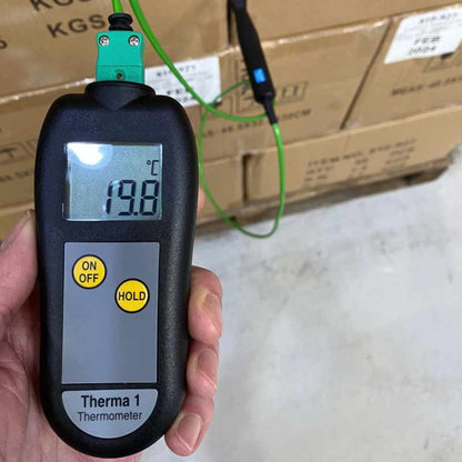 Une main tient un thermomètre industriel Therma 1 de Thermomètre.fr affichant une température de 19,8°C, avec un fil vert connecté à l'appareil, probablement une sonde à thermocouple. Les boîtes sont empilées en arrière-plan.